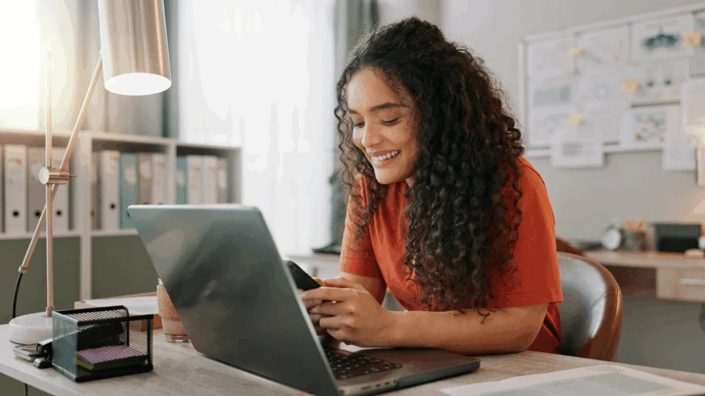 A woman with curly hair sits at a desk, focused on her laptop while working.