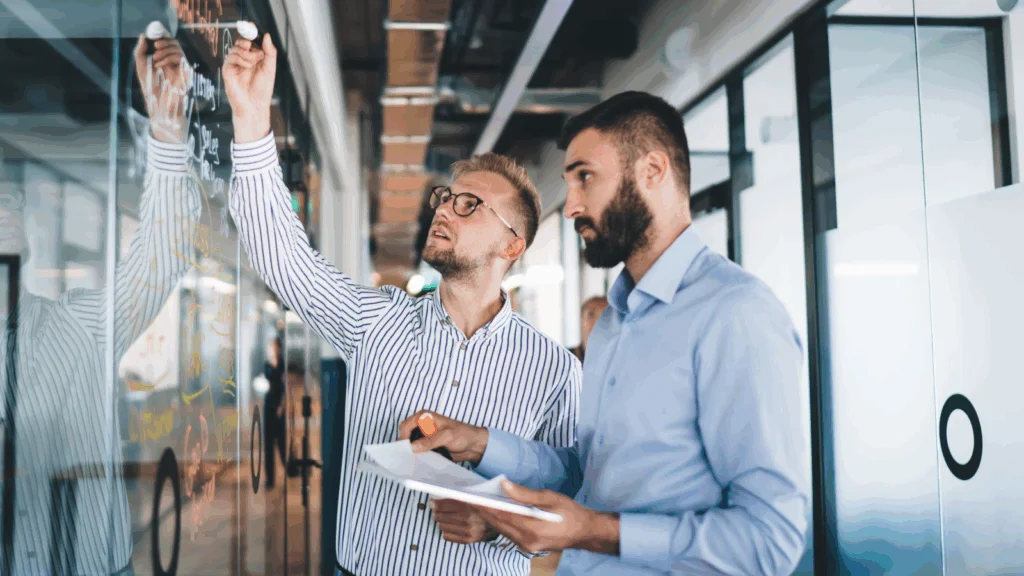 Two men standing side by side in front of a large glass wall, with reflections visible on the surface.