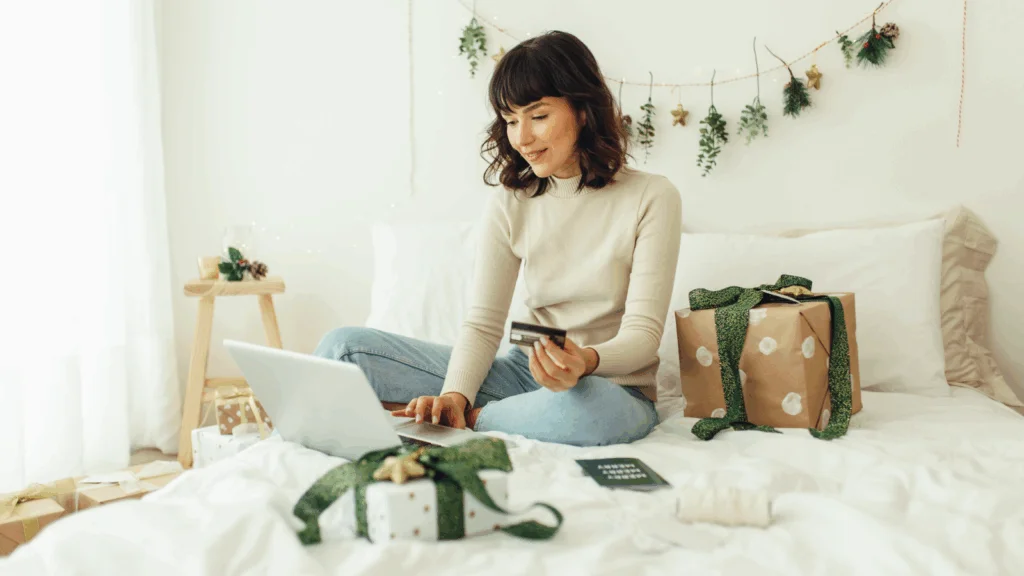 A woman is sitting on a bed and using a credit card to buy gifts online with Christmas decorations in the background.