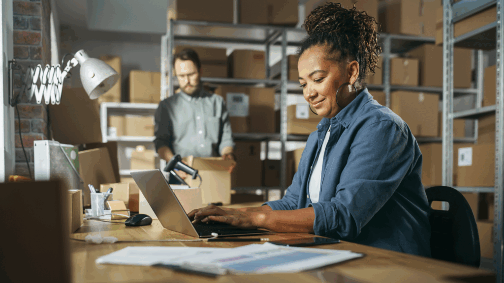 Why automating eBay fulfillment matters A woman focused on her laptop while seated in a busy warehouse filled with shelves and boxes.