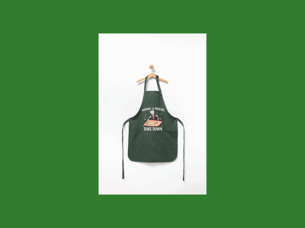 Green apron on a hanger against a white background, featuring a whimsical design with text "Having a Mental Bake Down”.