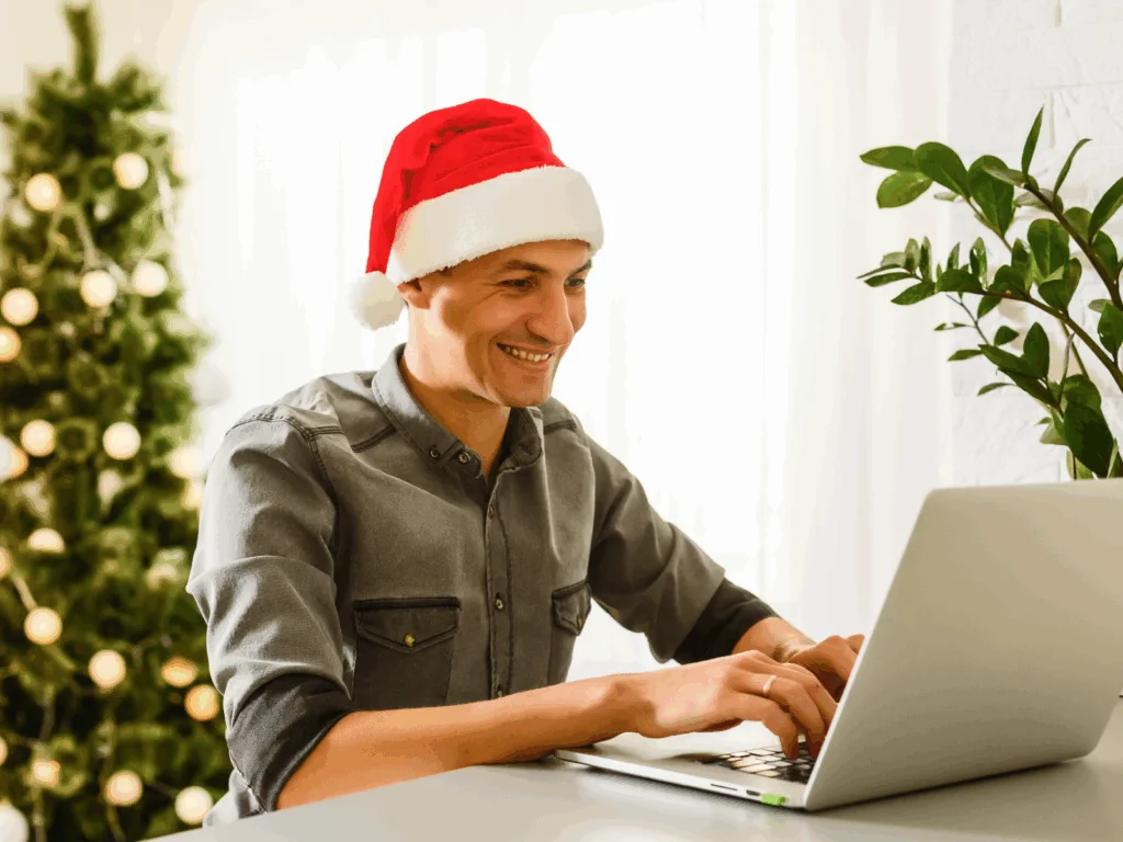 A smiling man wearing a Santa hat types on a laptop, searching for unique Christmas gifts, sitting indoors.