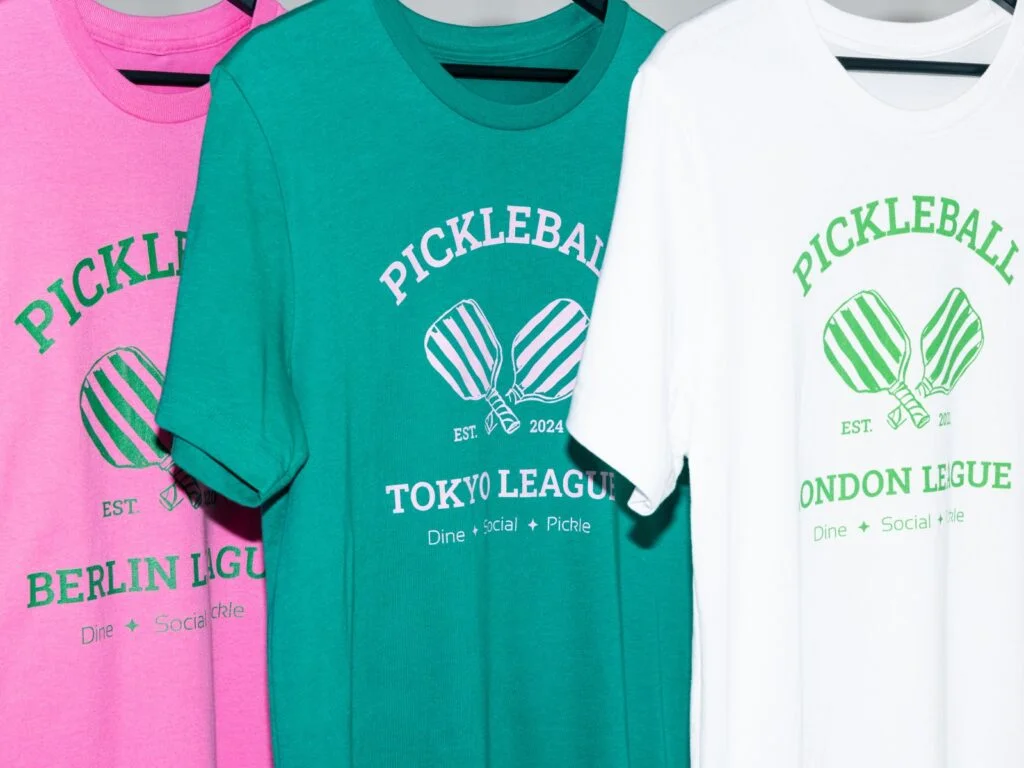 Pickleball t-shirts displayed on hangers, featuring a color scheme of white, pink, and green