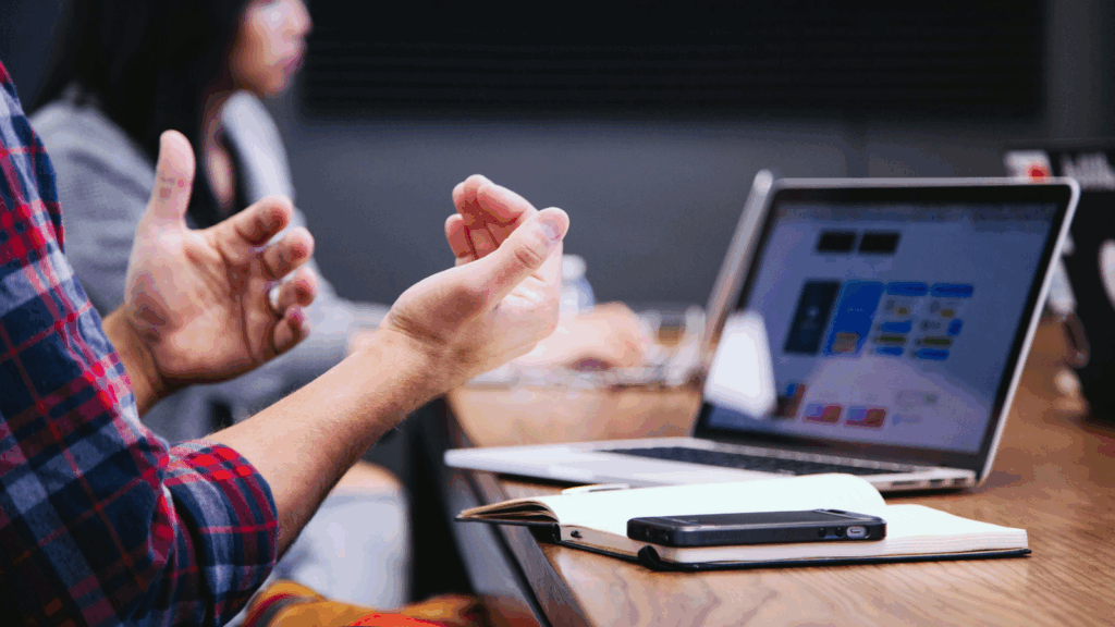 A man sitting at a desk with a laptop, gesturing as he speaks or explains something