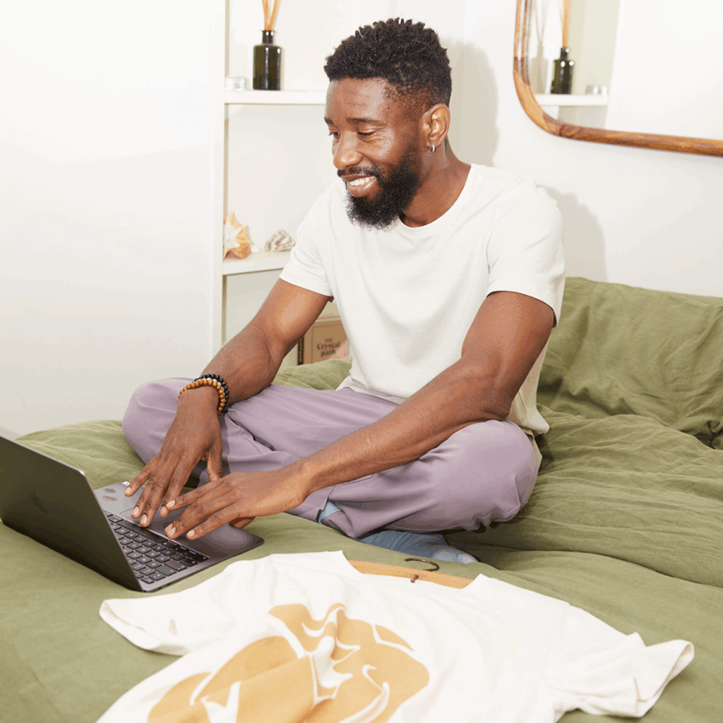 A man sitting cross-legged on a bed, with a t-shirt beside him, using a laptop to find the best shirts for sublimation printing
