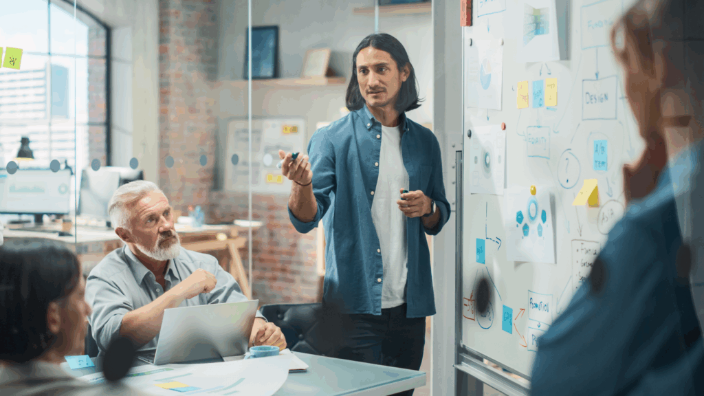 A man in a blue shirt presents ideas on a whiteboard to colleagues in a modern office.