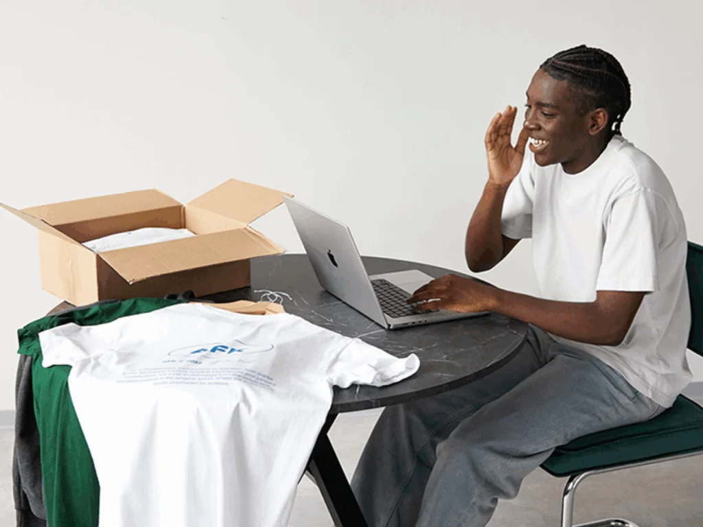 Common white label model challenges and how to overcome them A smiling young man in a white t-shirt is using a laptop while sitting at a table, with different clothes next to the laptop.