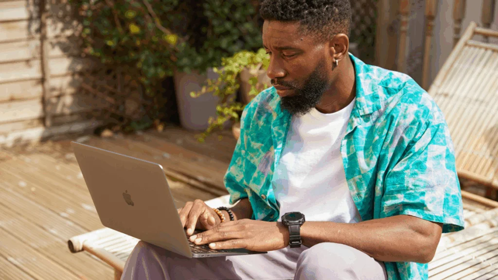 A man in a blue shirt sits outdoors on a wooden deck, using a laptop to find out how to make money on Canva.