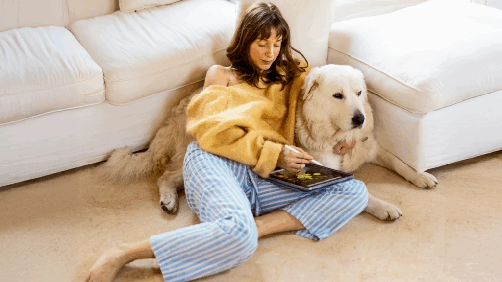 A woman in cozy striped pajamas and a yellow sweater sits on the floor, leaning against a white couch with a large dog by her side.