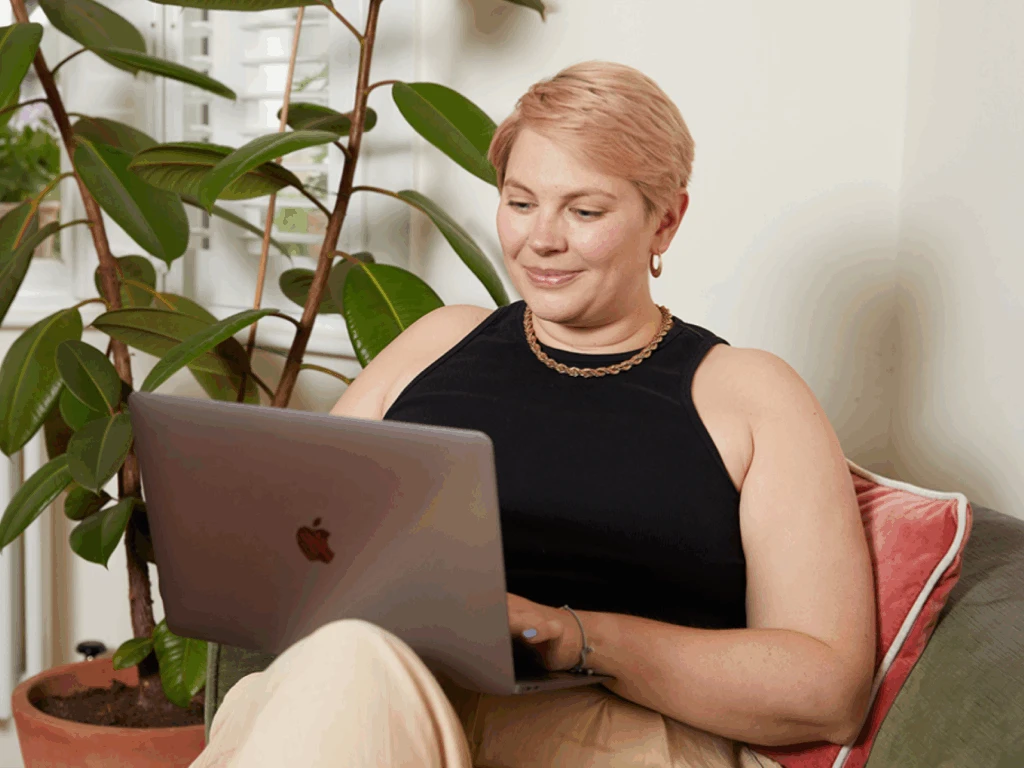 A woman with short hair is sitting on a couch, using a laptop, smiling and looking content.