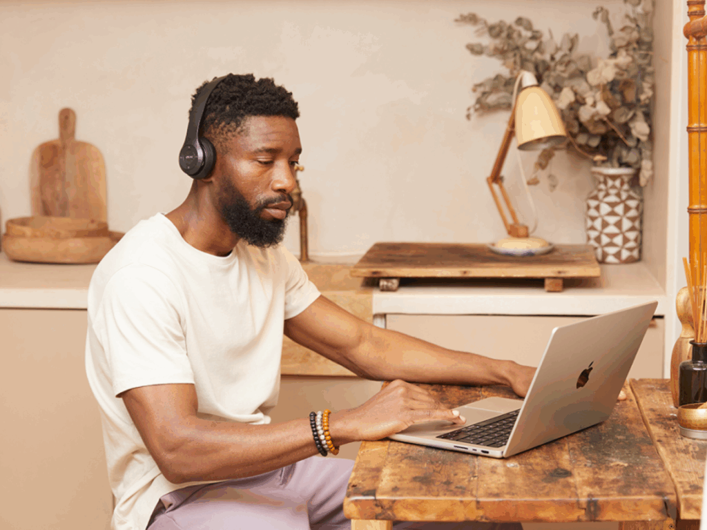 A man in a white t-shirt and headphones works on a laptop, designing gamer merch at a wooden table in a kitchen.