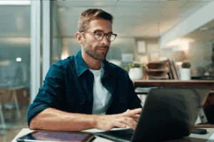 A man wearing glasses is focused on working on his laptop at a desk.
