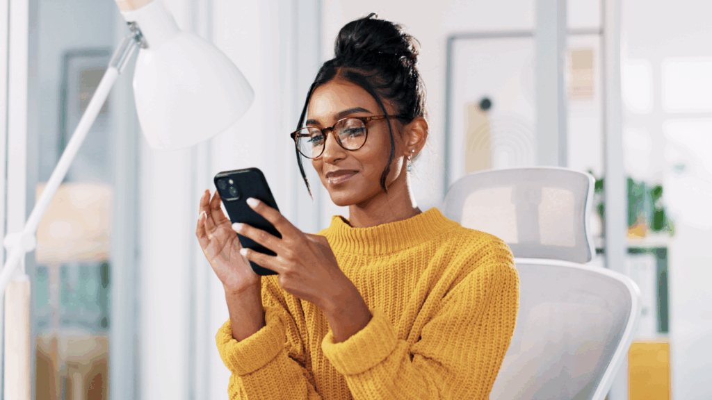 A woman in a yellow sweater and glasses smiles while looking at her phone in a modern office.
