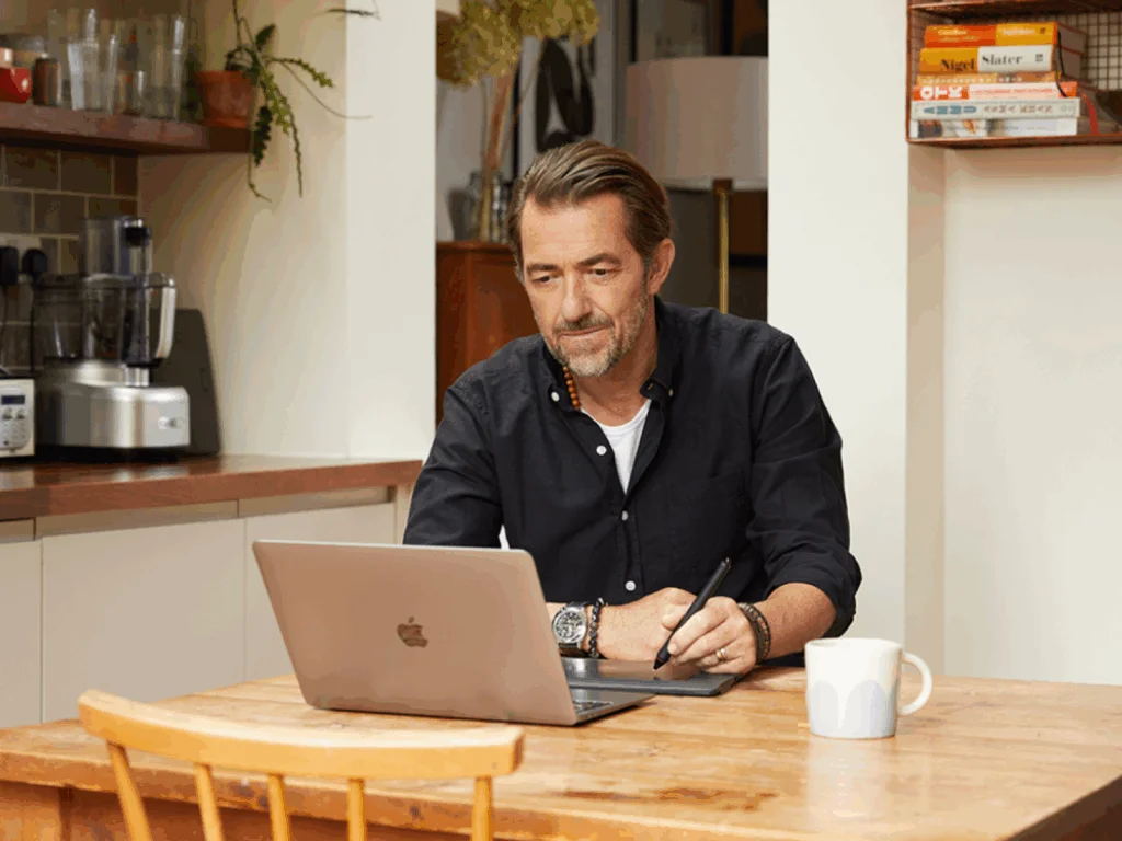 A man in a black shirt sits at a wooden table in a kitchen, working on a laptop while holding a stylus. A mug and books are nearby.