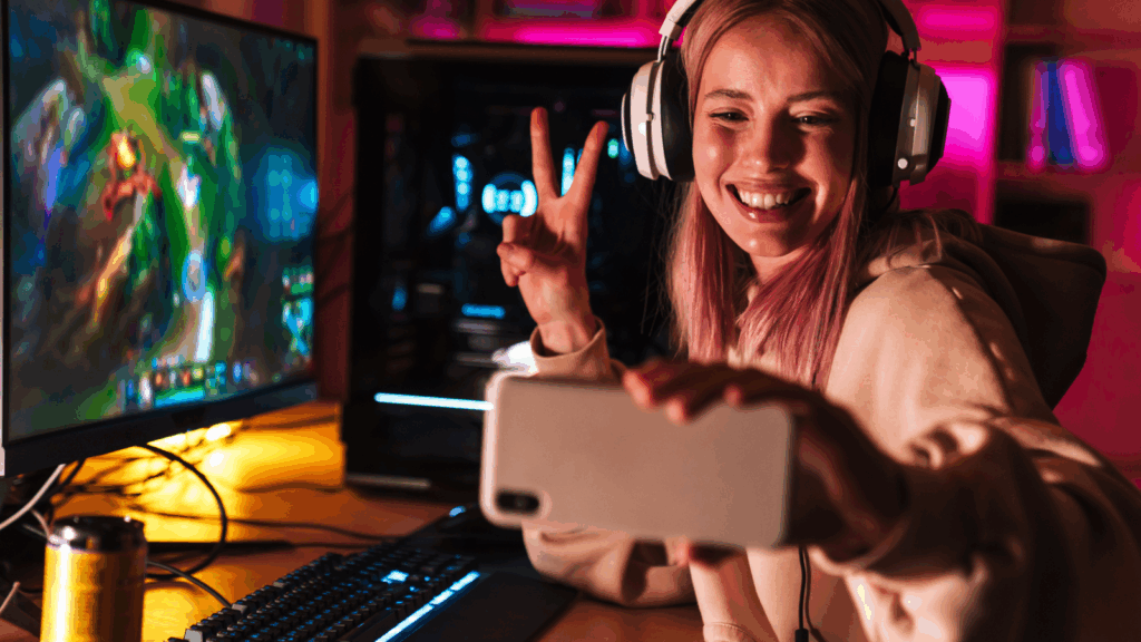 A woman with headphones, smiling and making a peace sign, takes a selfie at her gaming setup.