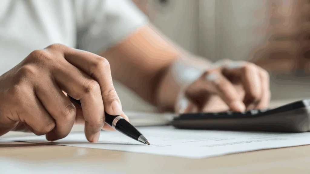 A person's hand writes on paper with a pen, while a calculator sits nearby, indicating a task involving calculations.