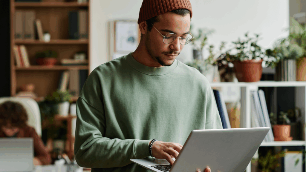 A man in a beanie and glasses types on a laptop, learning how to sell templates on Canva.