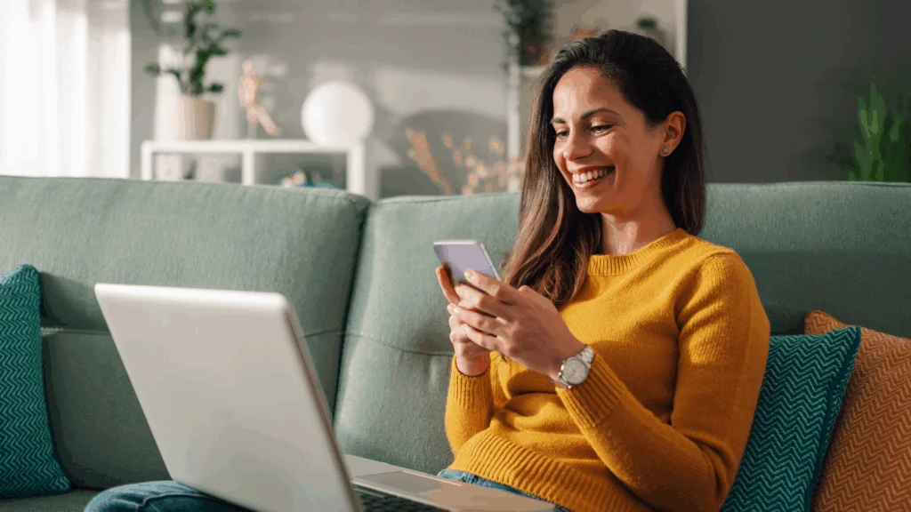 A woman in a yellow sweater sits on a green couch, smiling at her smartphone. An open laptop is on her lap.