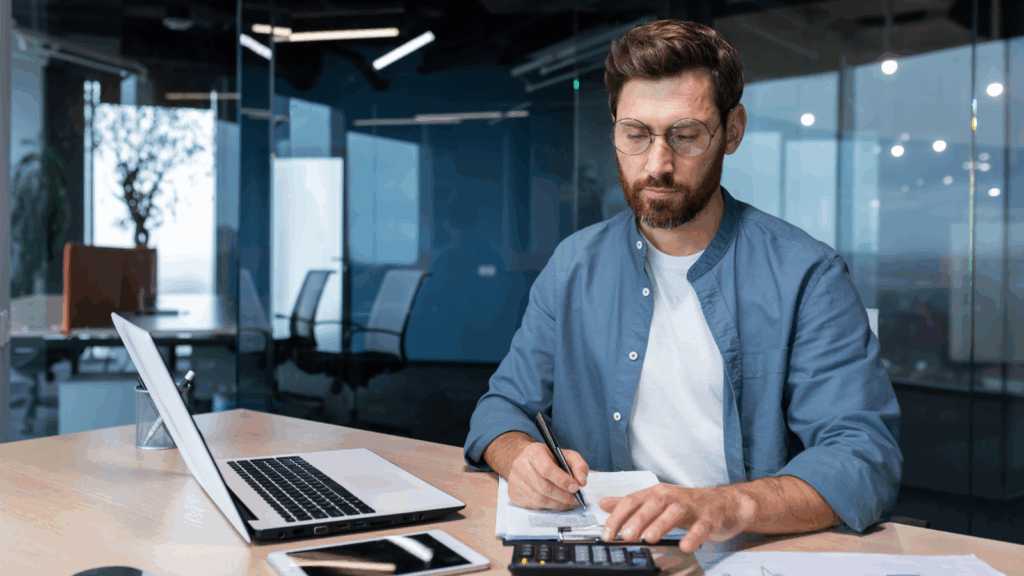 A man in a blue shirt works at a desk with a laptop, calculator, and notepad, focused and writing, in a glass-walled office.