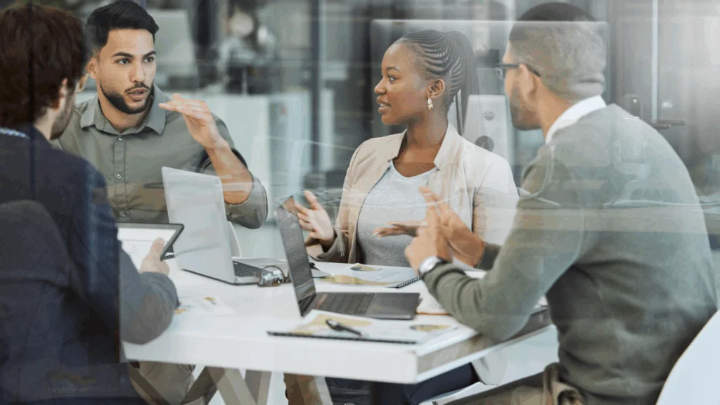 A group of four professionals engaged in a discussion around a table with laptops.