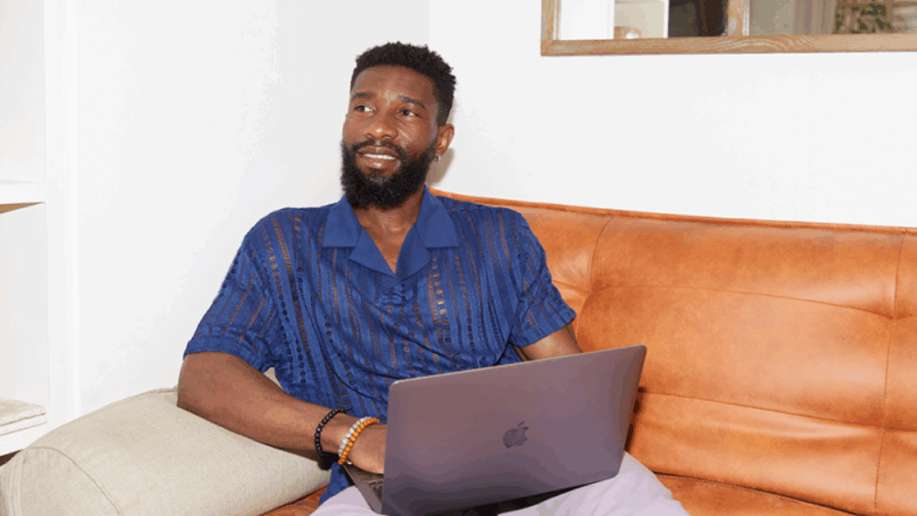 A man with a beard sits on a brown sofa, using a laptop to find out how much money do you need to start a business.