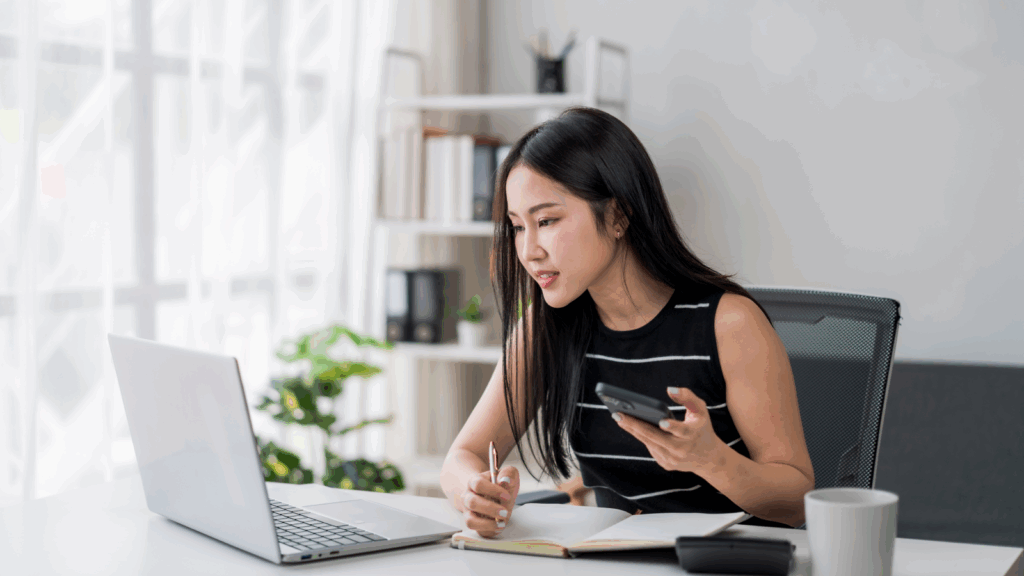 A woman is sitting at a desk, looking at a laptop while holding a phone, writing in a notebook.