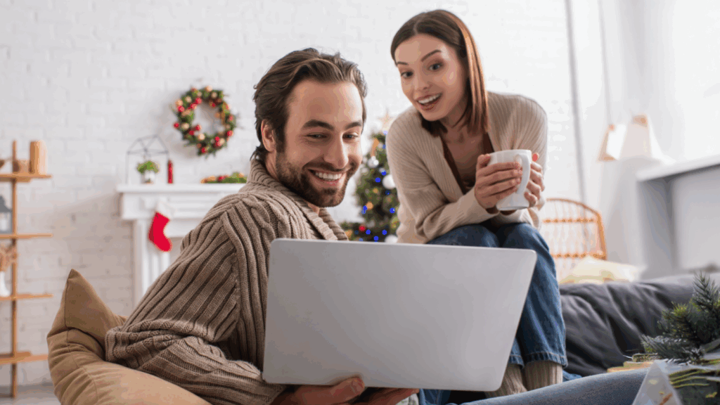 A smiling man and woman in cozy sweaters sit on a sofa, looking at a laptop, searching for DIY Christmas gift ideas.