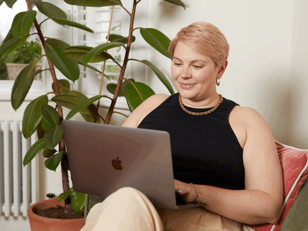 A woman with short hair sits comfortably on a couch, smiling while working on a laptop. A large houseplant is beside her.