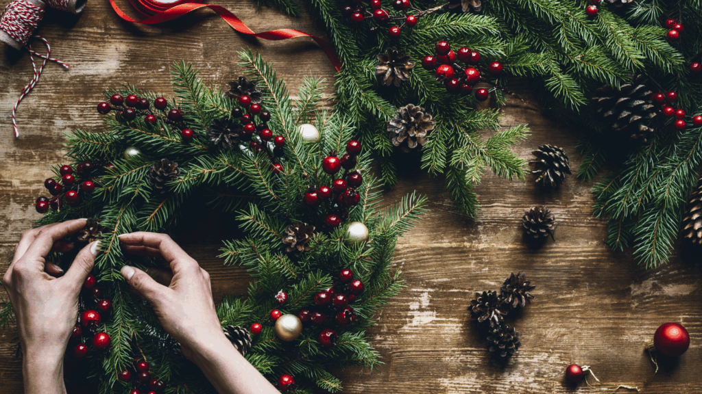 Hands assemble a festive Christmas wreath with green pine branches, red berries, pinecones, and ornaments.