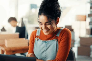 A smiling woman in an orange sweater and denim overalls uses a computer to find out if print-on-demand is worth it.