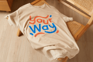 Beige t-shirt with "Your Way" in red and blue letters, draped over a woven wooden chair on a light wood floor.