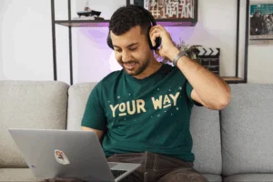 Smiling man in headphones, seated on a gray couch, using a laptop. Wears a green "Your Way" t-shirt.