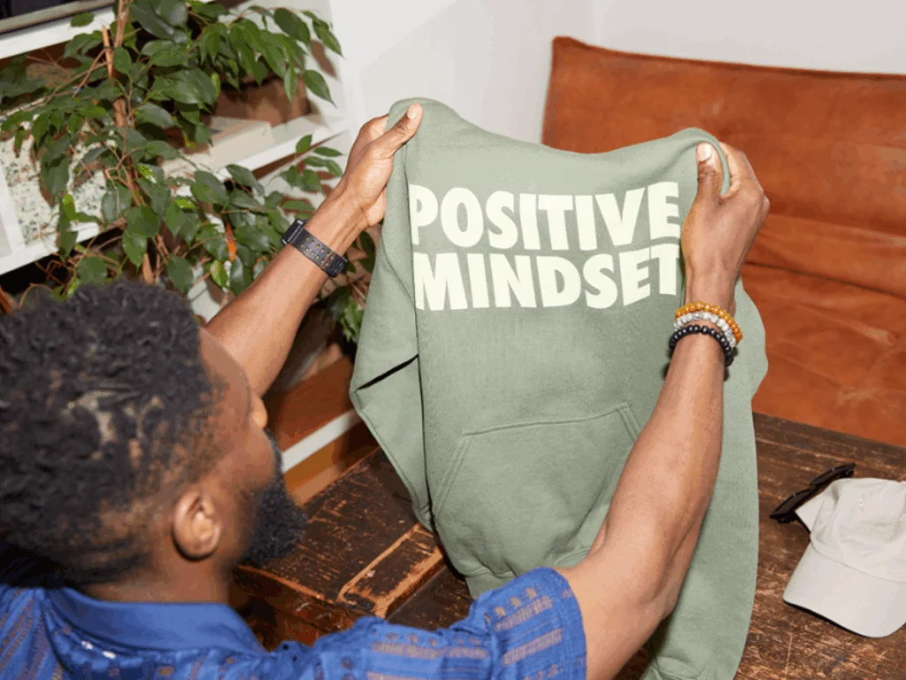 A man holds up a green hoodie with "Positive Mindset" in bold white letters.