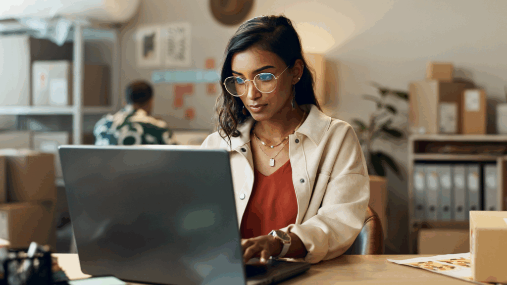 A focused woman in glasses works on a laptop in a cozy office with shelves of boxes.