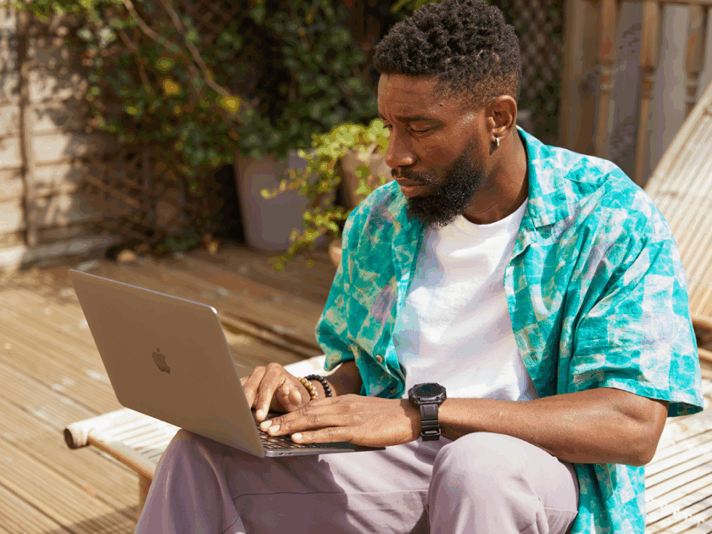 A man with a beard wears a colorful shirt and types on a laptop outdoors, searching for the most profitable POD products.