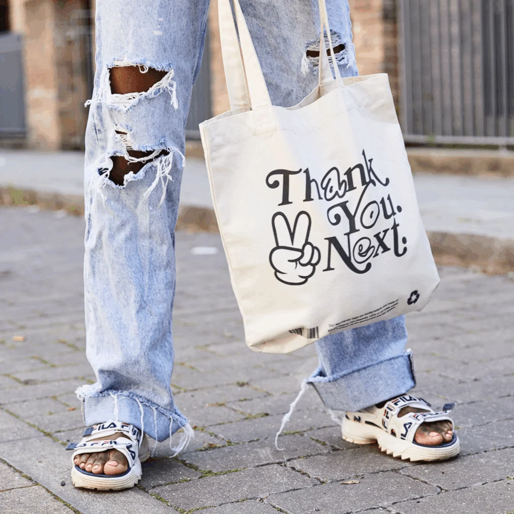 A woman in ripped jeans carries a tote bag that reads "thank you next," showcasing a casual and trendy style.
