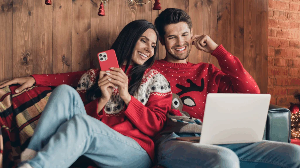 A cheerful couple in festive sweaters sits on a cozy couch, smiling at a laptop. One holds a smartphone.