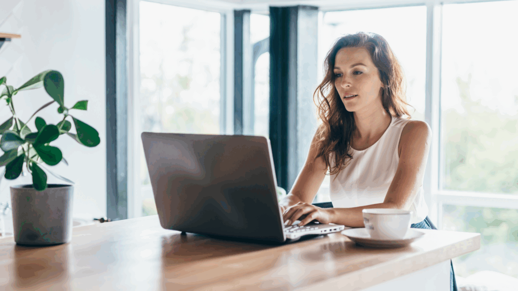 A woman in a white tank top works on a laptop at a wooden table. A potted plant and a cup of coffee are nearby.