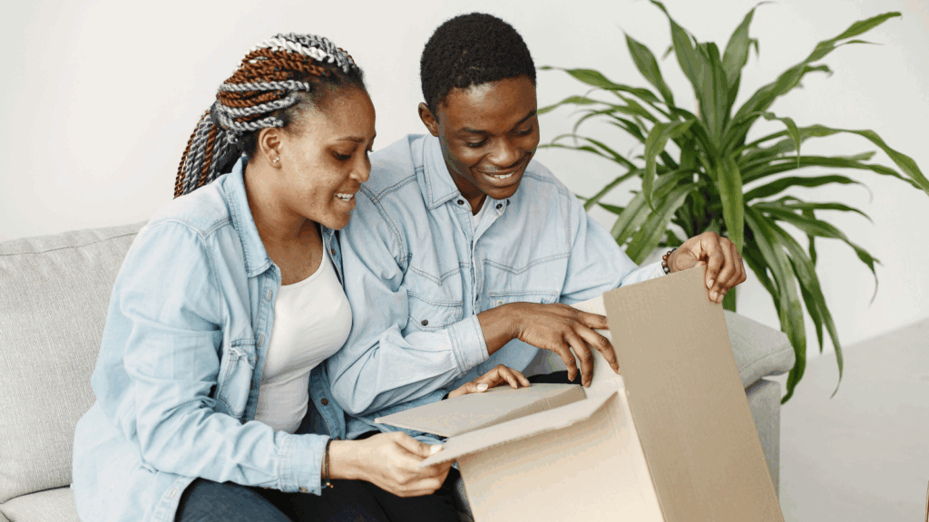 A young couple sits on a couch, excitedly opening a box together.
