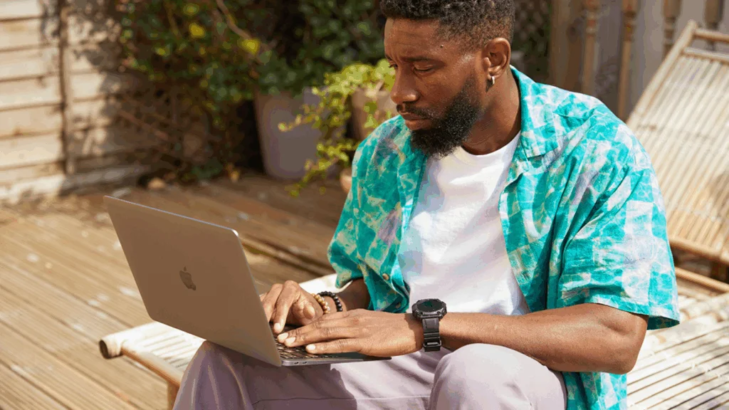 A man sitting on a wooden deck, focused on his laptop, with a serene outdoor setting in the background.