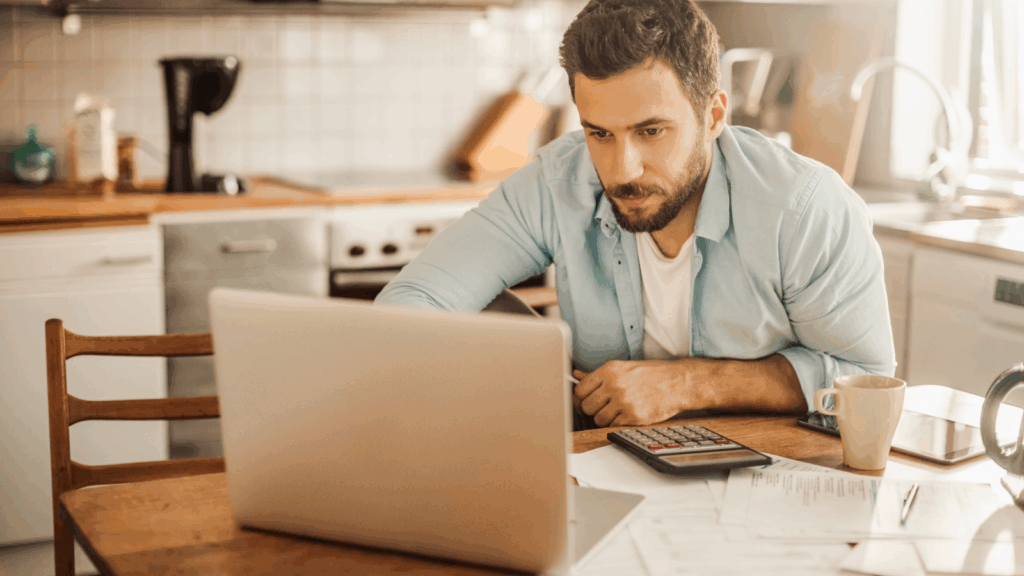 A man sits at a kitchen table, focused on a laptop. Papers, a calculator, and a mug are nearby.
