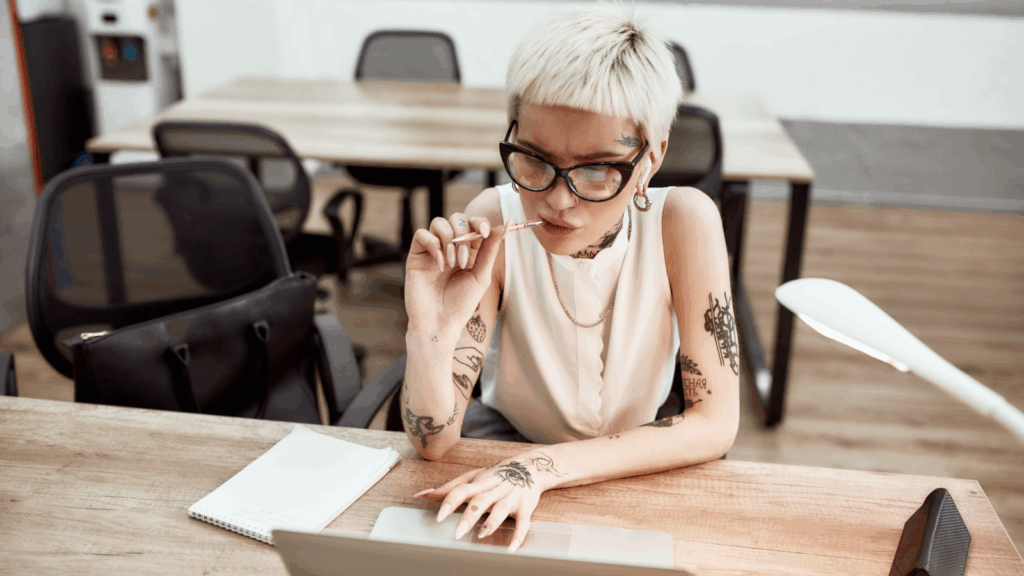 A focused person with short blonde hair and tattoos, wearing glasses, using a laptop to find out how to start a POD puzzle business.