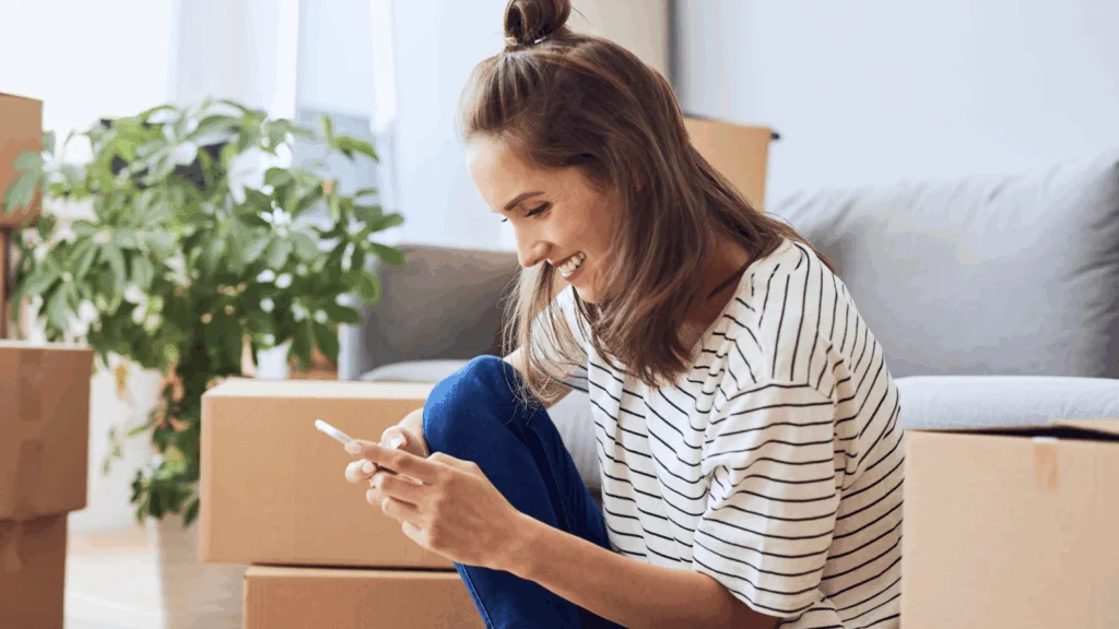 A woman in a striped shirt sits among moving boxes, smiling while looking at her phone, searching for a POD vs DIY comparison.