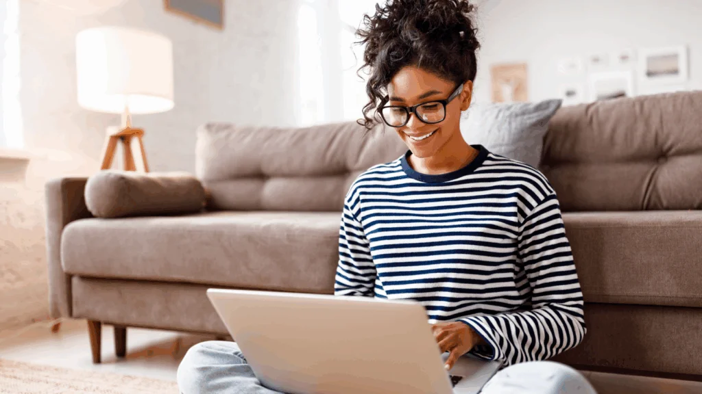A woman wearing glasses sits on the floor, focused on her laptop as she works or studies.