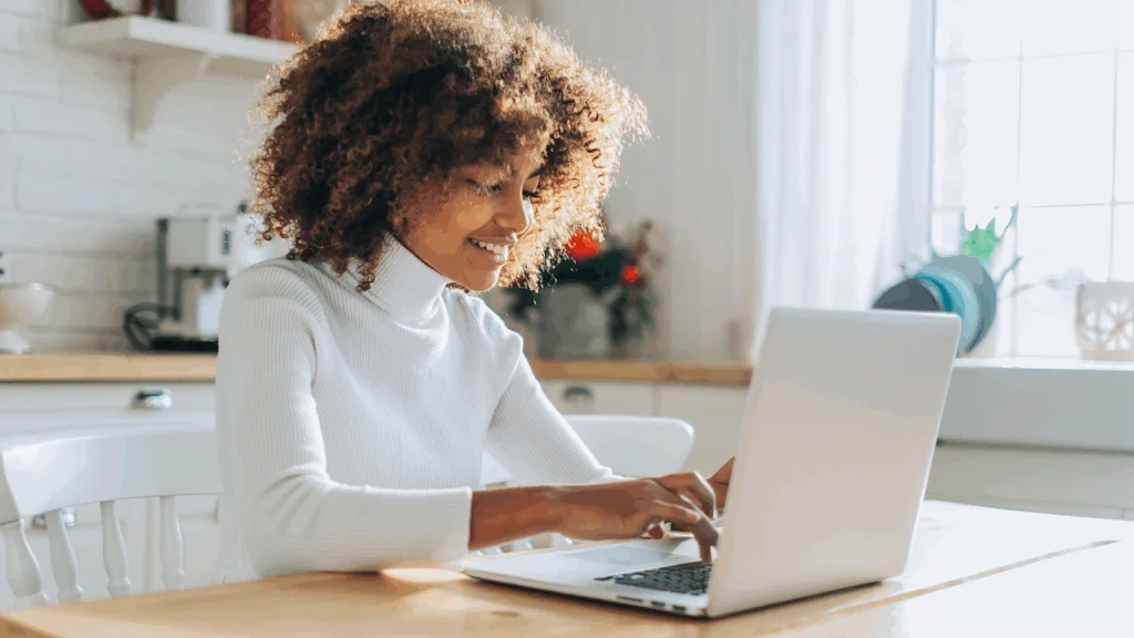 A woman with curly hair sits at a table, focused on her laptop, surrounded by a cozy indoor setting.