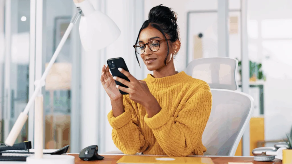 A woman in a yellow sweater, seated in a modern office, looks at her phone with a focused expression.