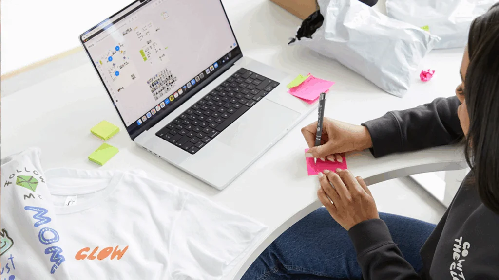 A woman writes on a pink sticky note at a desk with an open laptop, with a custom white t-shirt and packages nearby.