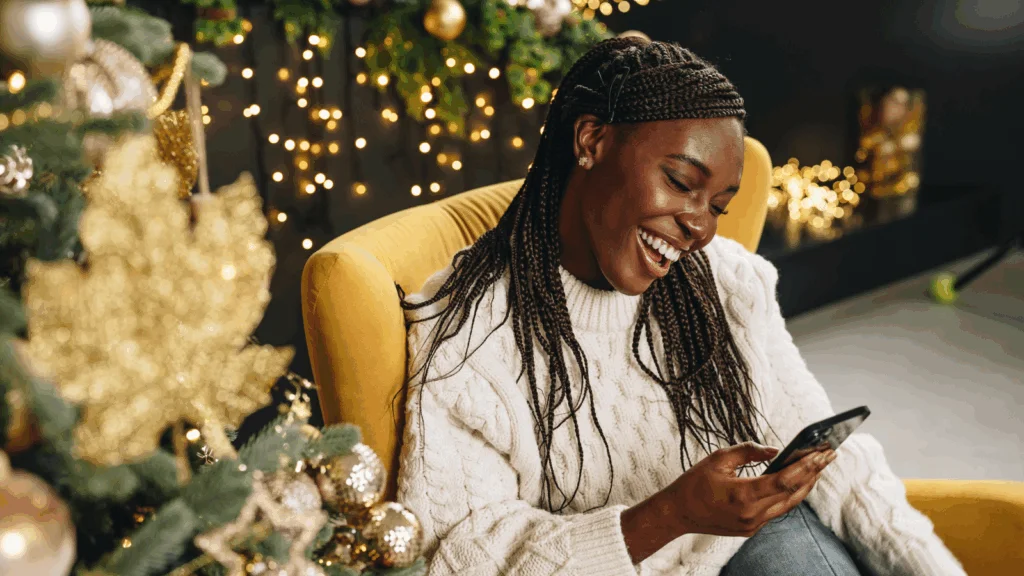 A woman in a cozy sweater laughs while looking at her phone, sitting on a yellow chair near a decorated Christmas tree.