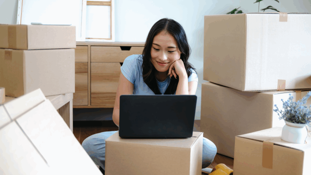 A woman sits on the floor surrounded by cardboard boxes, suggesting a move. She smiles while using a laptop.