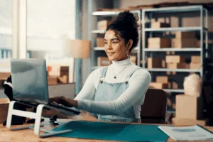 Smiling woman in a white turtleneck and denim overalls uses a laptop in a sunlit room with shelves and boxes.
